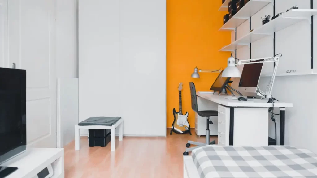 un appartement moderne en colocation. La pièce est lumineuse, avec des murs blancs et orange. Au premier plan, un lit soigneusement fait avec une couverture à carreaux est visible. 